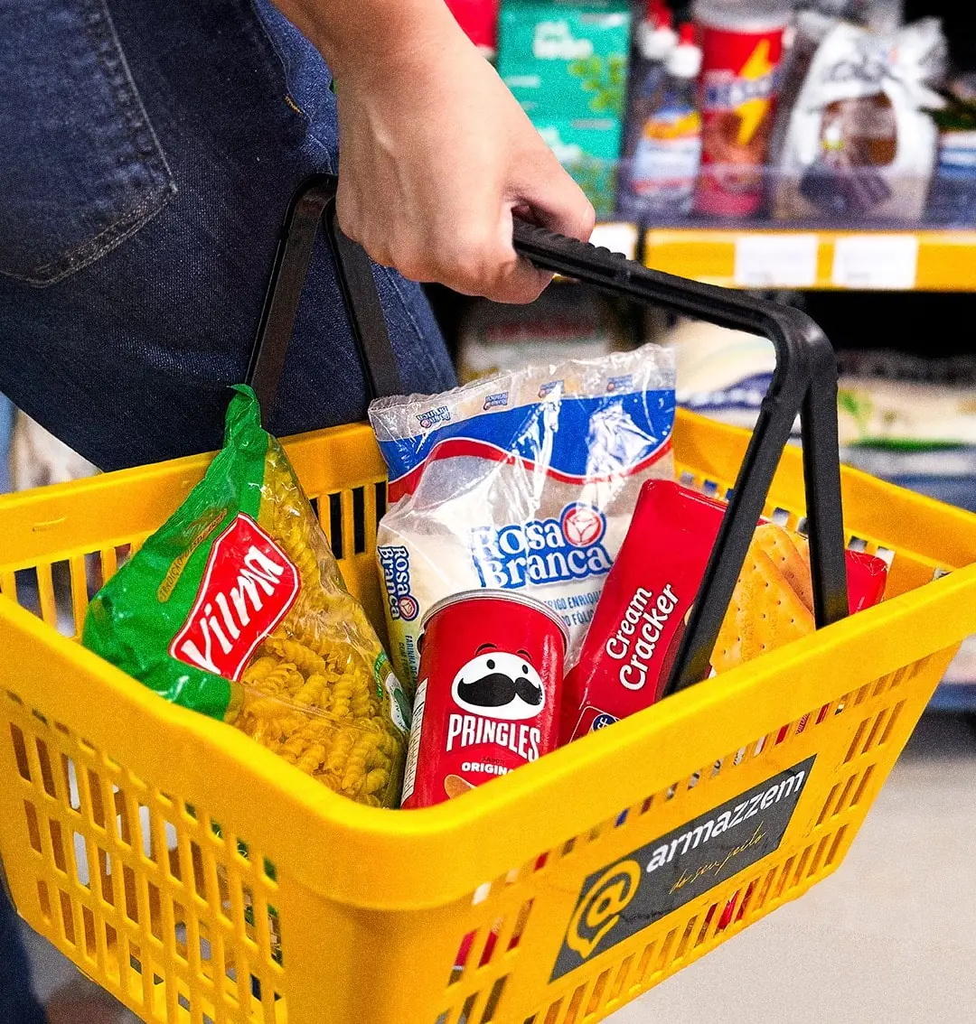Mão segurando a cesta amarela do Armazzem cheia de produtos