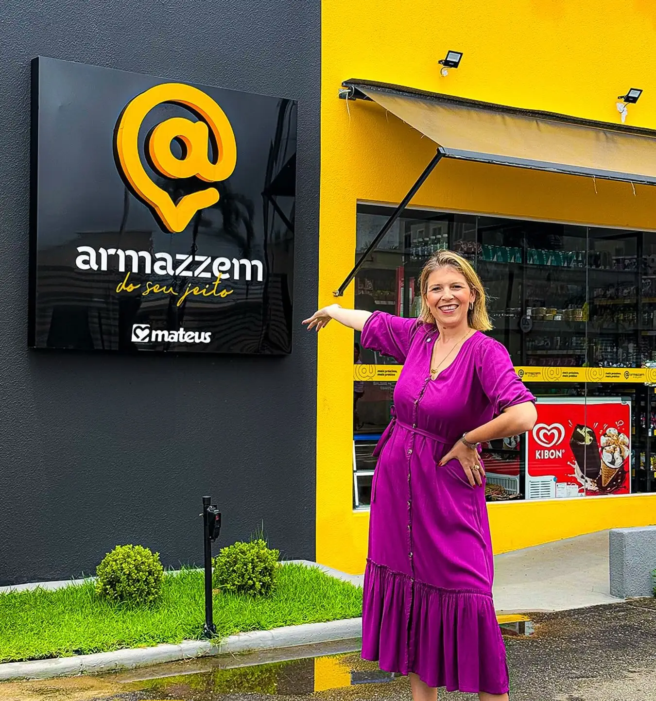 Mulher de vestido roxo apontando para a placa do Armazzem na fachada da loja